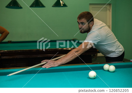 A man aims a cue at a billiard ball on a green table. A man hits a billiard ball and tries to pocket it. A man plays Russian billiards with his friends. Concept of a man playing billiards. A man aims a cue at a billiard ball on a green table. A man hits a billiard ball and tries to pocket it. A man plays Russian billiards with his friends. Concept of a man playing billiards. 120852205