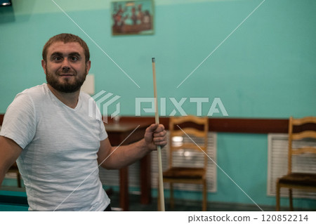 With a funny squinting of his eyes, a man stands with a cue, leaning on a billiard table, and looks into the camera. Copy space. A man jokes and makes faces in a billiard room, playing with friends. 120852214