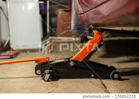The car is standing in the garage, raised with a jack, while a man is repairing it Close-up of a jack under the bottom of a car in a garage. The jack holds the car while the person changes the wheel. 120852218