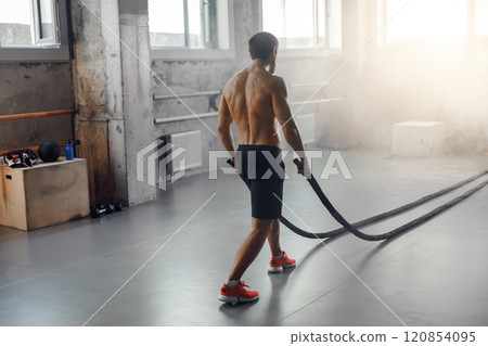 Fitness Trainer Engaged in Exercising with Battle Ropes in a Dynamic Gym Environment Fitness Trainer Engaged in Exercising with Battle Ropes in a Dynamic Gym Environment 120854095