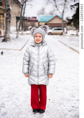 8-year-old Russian girl smiles in the snow, wearing a warm winter jacket and hat. She stands outdoors in a snowy landscape, looking at the camera with a joyful, playful expression 120855403
