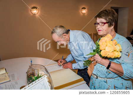 An elderly couple signs documents at a registry office in Russia, celebrating their 50th wedding anniversary. The woman holds a bouquet of yellow roses, symbolizing love and commitment 120855605