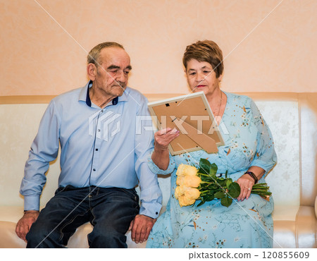 76-year-old man and 71-year-old woman celebrate their 50th wedding anniversary. The woman in a blue dress holds yellow roses, and they look at a gift a frame with a diploma and photo 76-year-old man and 71-year-old woman celebrate their 50th wedding anniversary. The woman in a blue dress holds yellow roses, and they look at a gift a frame with a diploma and photo 120855609