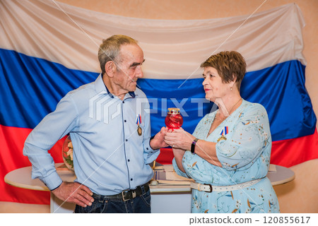 76-year-old man and a 71-year-old woman celebrate their golden wedding anniversary (50 years) in festive attire, holding a red lamp symbolizing faith and love, with medals, against the Russian flag 120855617