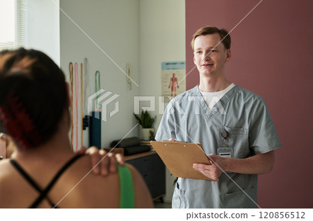Smiling Doctor Holding Clipboard in Examination Room 120856512