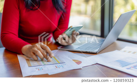 Woman wear a red shirt  holding a pen and smartphone while working at office, Close up shot hands of woman use pen to analyze graphs and business data. 120859323