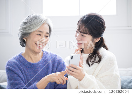 Close-up of mother and daughter smiling and talking in the living room while looking at their smartphones Close-up of mother and daughter smiling and talking in the living room while looking at their smartphones 120862604
