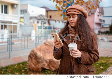 Mid shot of a young Caucasian woman in a beret and coat using smartphone on the street and holds cup of coffee. Concept of modern social media 120864362