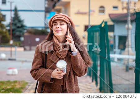 Portrait of a young Caucasian woman in a beret and coat smiling, calling at smartphone on the street and holds cup of coffee. Concept of modern social media 120864373