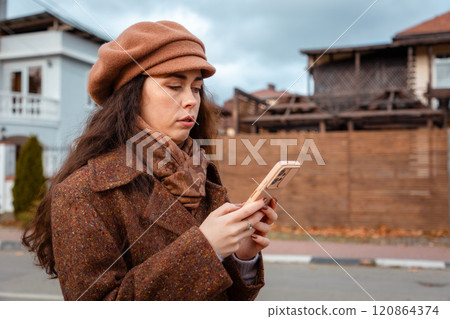 Portrait of a young Caucasian woman in a beret and coat using a smartphone on the street. Concept of modern social media and online communication 120864374
