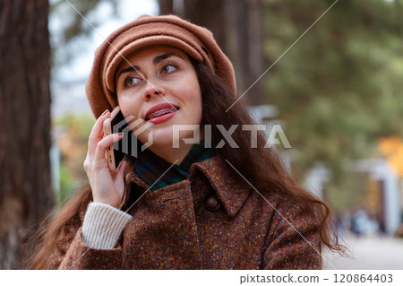 Portrait of a young pretty Caucasian woman in a hat and coat smiling and talking on phone. Modern communication and business 120864403