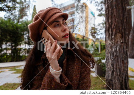 Side view of a young pretty Caucasian woman in a hat and coat talking on phone. Modern communication and business Side view of a young pretty Caucasian woman in a hat and coat talking on phone. Modern communication and business 120864405