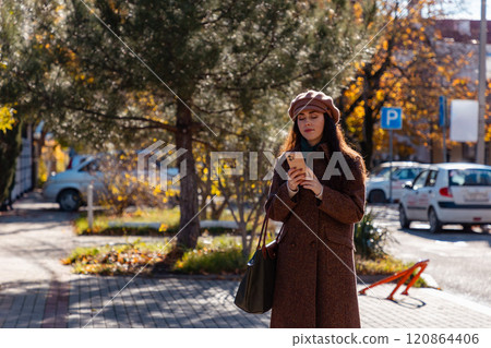 Wide shot of a young Caucasian woman in a autumn coat and beret using smartphone and walking on autumn street. Modern communication and business Wide shot of a young Caucasian woman in a autumn coat and beret using smartphone and walking on autumn street. Modern communication and business 120864406