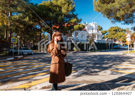 Wide shot of a young pretty Caucasian woman in a hat and coat talking on phone. Concept of modern communication and business Wide shot of a young pretty Caucasian woman in a hat and coat talking on phone. Concept of modern communication and business 120864407