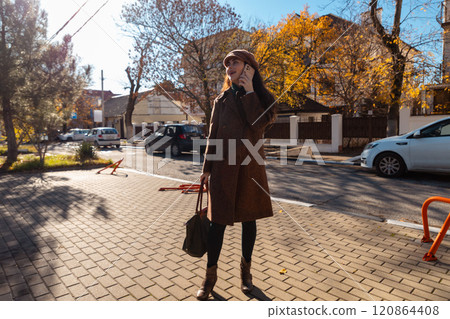 Wide view of a Caucasian stylish young woman in a coat and beret, smiling and calling on a smartphone on the street. City lifestyle Wide view of a Caucasian stylish young woman in a coat and beret, smiling and calling on a smartphone on the street. City lifestyle 120864408