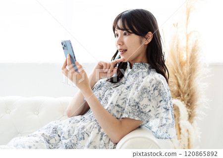 A young woman with long black hair sitting on a white sofa and holding a smartphone 120865892