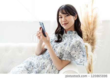 A young woman with long black hair sitting on a white sofa and holding a smartphone A young woman with long black hair sitting on a white sofa and holding a smartphone 120865896