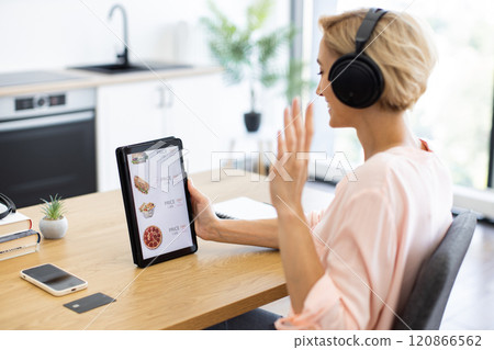 Happy Caucasian woman using tablet to order food online while enjoying music in headphones. Relaxed atmosphere with modern kitchen background, highlighting ease of online shopping at home. 120866562