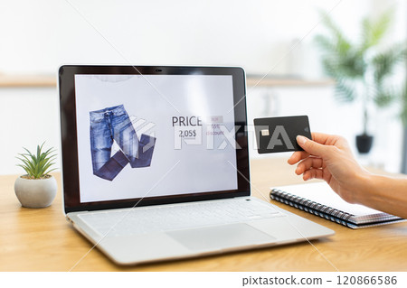 Caucasian woman shopping online using credit card and laptop for payment. She browses clothing options on digital platform, demonstrating virtual shopping experience. 120866586
