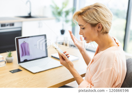 Caucasian adult woman expressing frustration while making phone call to discuss online clothing order issue. Her laptop shows purchased item confirming concern. Caucasian adult woman expressing frustration while making phone call to discuss online clothing order issue. Her laptop shows purchased item confirming concern. 120866596