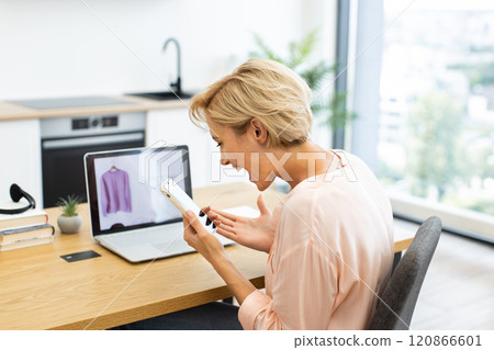 Caucasian woman expressing frustration while making phone call about online shopping problem. She sits at desk with laptop showing clothing item. Home office implies contemporary shopping experience. 120866601