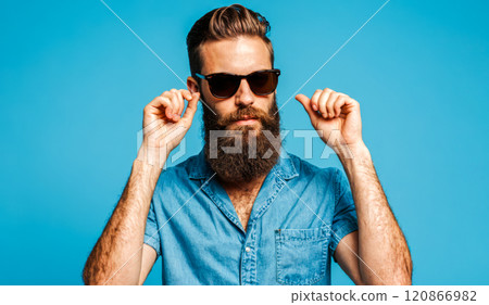 Stylish bearded man sporting sunglasses denim attire vibrant blue background. urban chic summer fashion casual fashionable male figure trendy beard cool posture natural light 120866982
