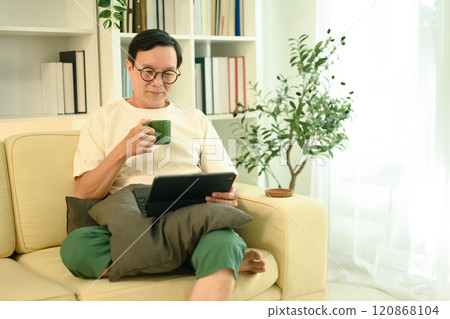 Happy middle aged man sipping coffee from a green mug and browsing on digital tablet Happy middle aged man sipping coffee from a green mug and browsing on digital tablet 120868104