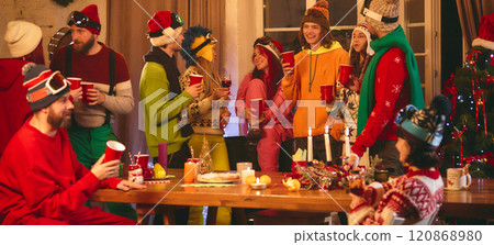 Warm holiday party scene as friends in ski-themed attire share laughter and drinks around wooden table glowing with festive candles and seasonal treats. Warm holiday party scene as friends in ski-themed attire share laughter and drinks around wooden table glowing with festive candles and seasonal treats. 120868980