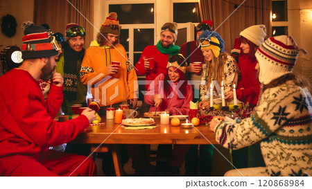 Holiday cheer abounds as friends in vibrant ski gear toast with red cups around candlelit table, sharing pastries, oranges, and laughter in warm setting. 120868984