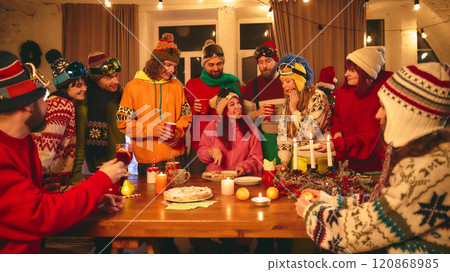 Festive holiday celebration with friends in ski-themed winter outfits gathered around wooden table, toasting with drinks and enjoying pastries by candlelight. Festive holiday celebration with friends in ski-themed winter outfits gathered around wooden table, toasting with drinks and enjoying pastries by candlelight. 120868985
