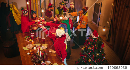 Joyful group in ski gear gathers in festive room, raising red cups and sharing snacks around wooden table lit by candles and surrounded by glowing lights. 120868986