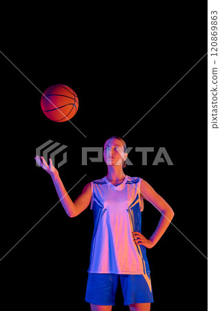 Portrait of young girl in uniform, basketball player bouncing ball on hand against black studio background in neon light 120869863