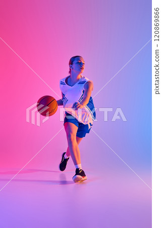 Full-length dynamic image of teen girl in uniform, basketball player in motion with ball during training session against gradient pink blue background in neon light 120869866