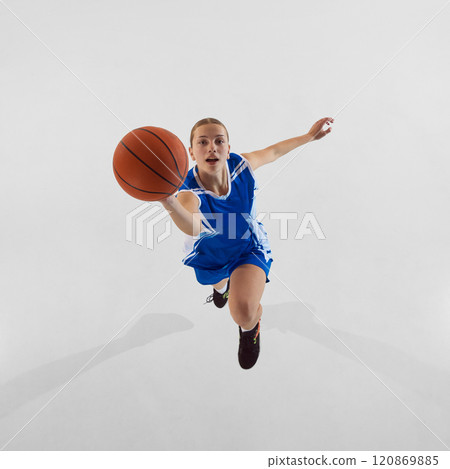 Slam dunk. Top view dynamic image of young focused teen girl in blue uniform, basketball player in motion, jumping with ball against white studio background 120869885