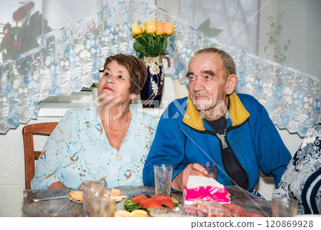 A Russian couple, both over 70 years old, look to the side at a television. They are dressed in festive attire, with glasses on the holiday table in the background 120869928