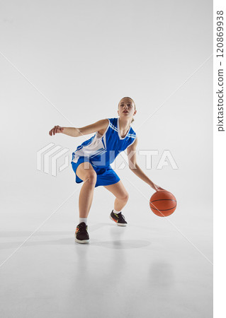 Young motivated and focused teen girl, basketball player in blue uniform, with ball, training against white studio background 120869938