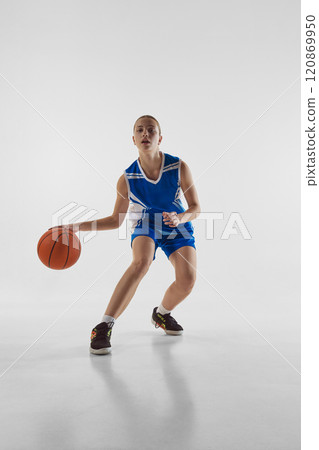 Dribbling. Full-length image of young concentrated teen girl in blue uniform, playing basketball, training against white studio background Dribbling. Full-length image of young concentrated teen girl in blue uniform, playing basketball, training against white studio background 120869950