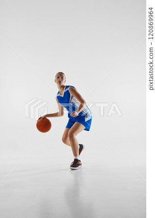 Dribbling. Full-length image of young concentrated teen girl in blue uniform, playing basketball, training against white studio background 120869964