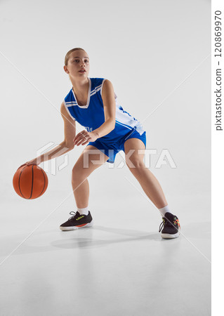 Concentrated young teen girl, basketball player in blue uniform in motion with ball during game, training against white studio background Concentrated young teen girl, basketball player in blue uniform in motion with ball during game, training against white studio background 120869970