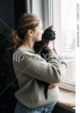 Pregnant woman holding black cat by the window, questioning the safety of keeping pets during pregnancy. Cozy home with coffee and candle. 120871159