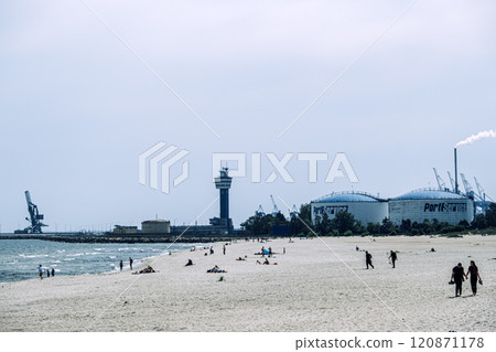 Beach with people relaxing near an industrial port and control tower on a cloudy day. Contrast of leisure and industry. Gdansk, Poland - May 19, 2024 120871178