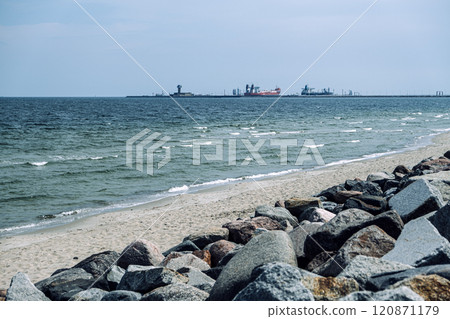 Rocky shoreline with waves rolling onto sandy beach and distant view of industrial ships and control tower. Coastal scene. Gdansk, Poland - May 19, 2024 120871179