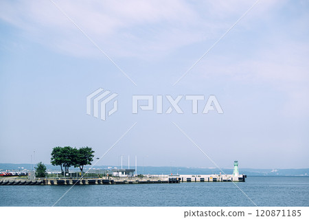 Minimalistic pier with a green lighthouse and trees surrounded by calm waters under a hazy sky. Tranquil coastal scene. Gdansk, Poland - May 19, 2024 120871185