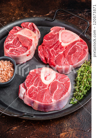 Raw Osso buco Veal shank steak, meat Ossobuco in steel tray. Dark background. Top view Raw Osso buco Veal shank steak, meat Ossobuco in steel tray. Dark background. Top view 120873871
