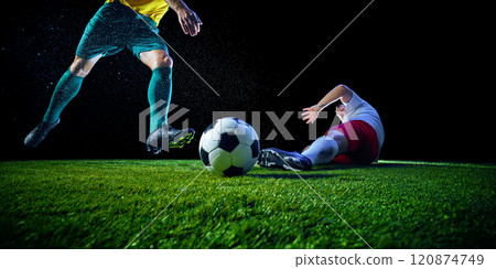 Soccer player in green kicks ball fiercely, while on green football field while other team's defender tries to stop him against black studio background. 120874749