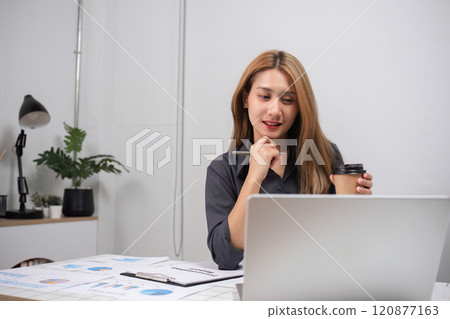 Young Professional Woman Analyzing Work on Laptop in Modern Office Environment with Coffee and Documents 120877163