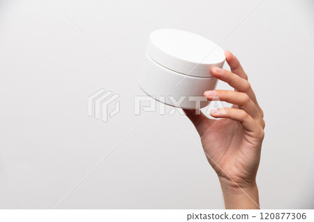 Close-up of a woman's hand holding a white jar of cream, mockup for advertising. Girl on a white background showing a cosmetic product Close-up of a woman's hand holding a white jar of cream, mockup for advertising. Girl on a white background showing a cosmetic product 120877306