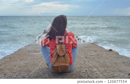 Rear view of a young woman tourist with a backpack sitting on a pier and waves hitting the pier in a storm. City of Sudak, Crimea 120877554