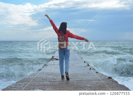 Rear view of a young woman tourist with a backpack standing on the pier and waves hitting the pier in a storm. City of Sudak, Crimea Rear view of a young woman tourist with a backpack standing on the pier and waves hitting the pier in a storm. City of Sudak, Crimea 120877555