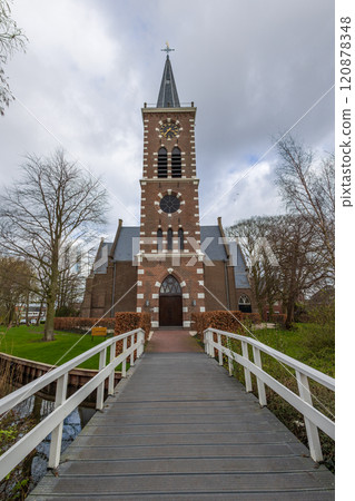 A picturesque brick church with a tall spire, surrounded by bare trees, viewed from a wooden footbridge. The cloudy sky adds depth to this serene and timeless scene. Church near Amsterdam. A picturesque brick church with a tall spire, surrounded by bare trees, viewed from a wooden footbridge. The cloudy sky adds depth to this serene and timeless scene. Church near Amsterdam. 120878348
